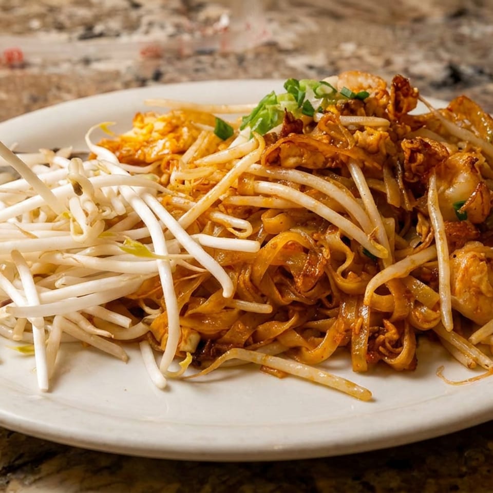 A white plate of Pad Thai noodles with shrimp, bean sprouts, and green onions on a marble table.
